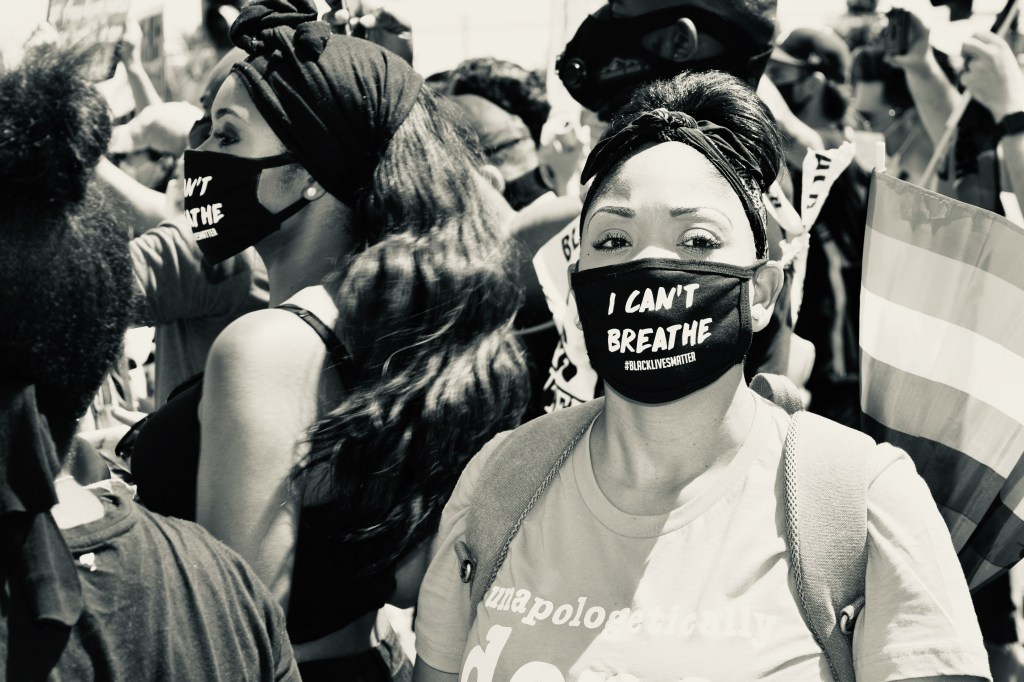 A woman in a crowd wearing a mask with the words "I can't breath, #Blacklivesmatter" on it.