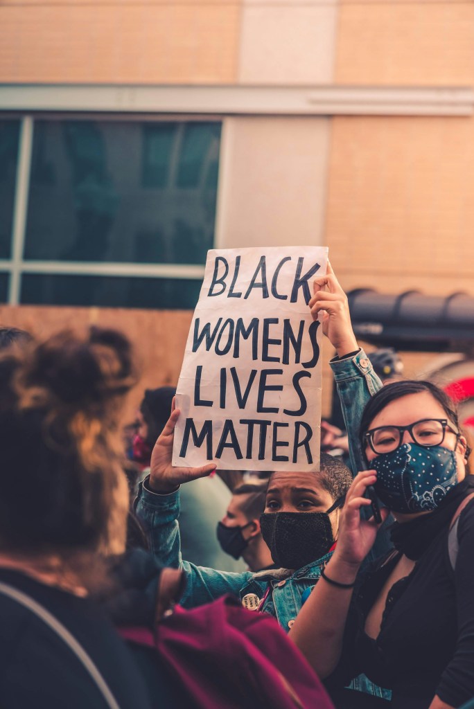 A masked women in a crowd holding up a sign that says "Black Womens Lives Matter."